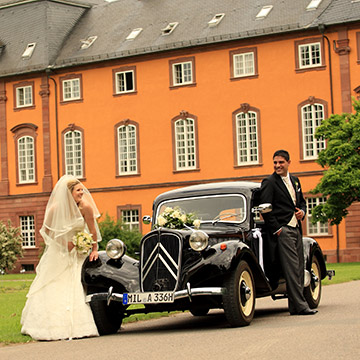 Hochzeit in Amorbach. Das Brautpaar neben Oldtimer vorm Schloss.